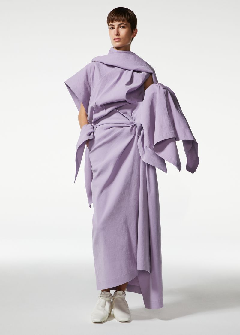 Fashion shot of a woman in a lilac dress with large draped details and white shoes, posing in a brightly lit studio against a white backdrop.