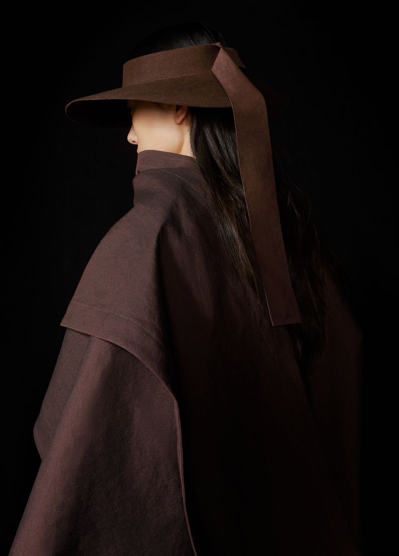 A woman wearing a brown hat with a wide brim and ribbon detail and a brown jacket against a dark background in a studio setting.