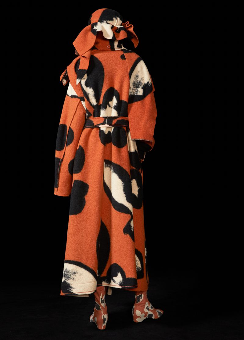 Person wearing an orange coat with black and white prints and matching boots and a hood in a studio setting with a dark background.