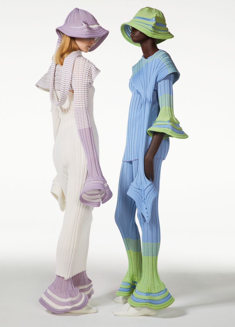 Two models showcase knitted fashion with matching bucket hats in a light studio setting. The outfits feature unique, avant-garde designs.