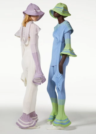 Two models showcase knitted fashion with matching bucket hats in a light studio setting. The outfits feature unique, avant-garde designs.