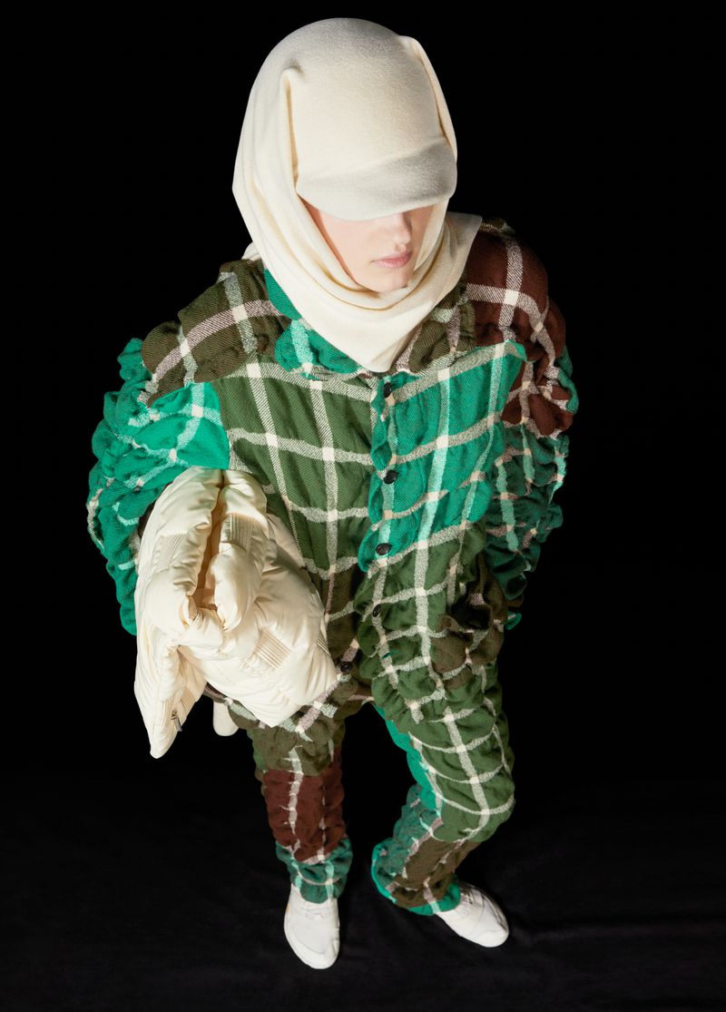 A person wears a green and brown plaid jumpsuit and a white head covering, posed in a studio with a dark background, top-down shot.