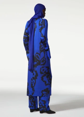 Model from behind in a vibrant royal blue and black floral coat, matching trousers, and head covering on a white backdrop.