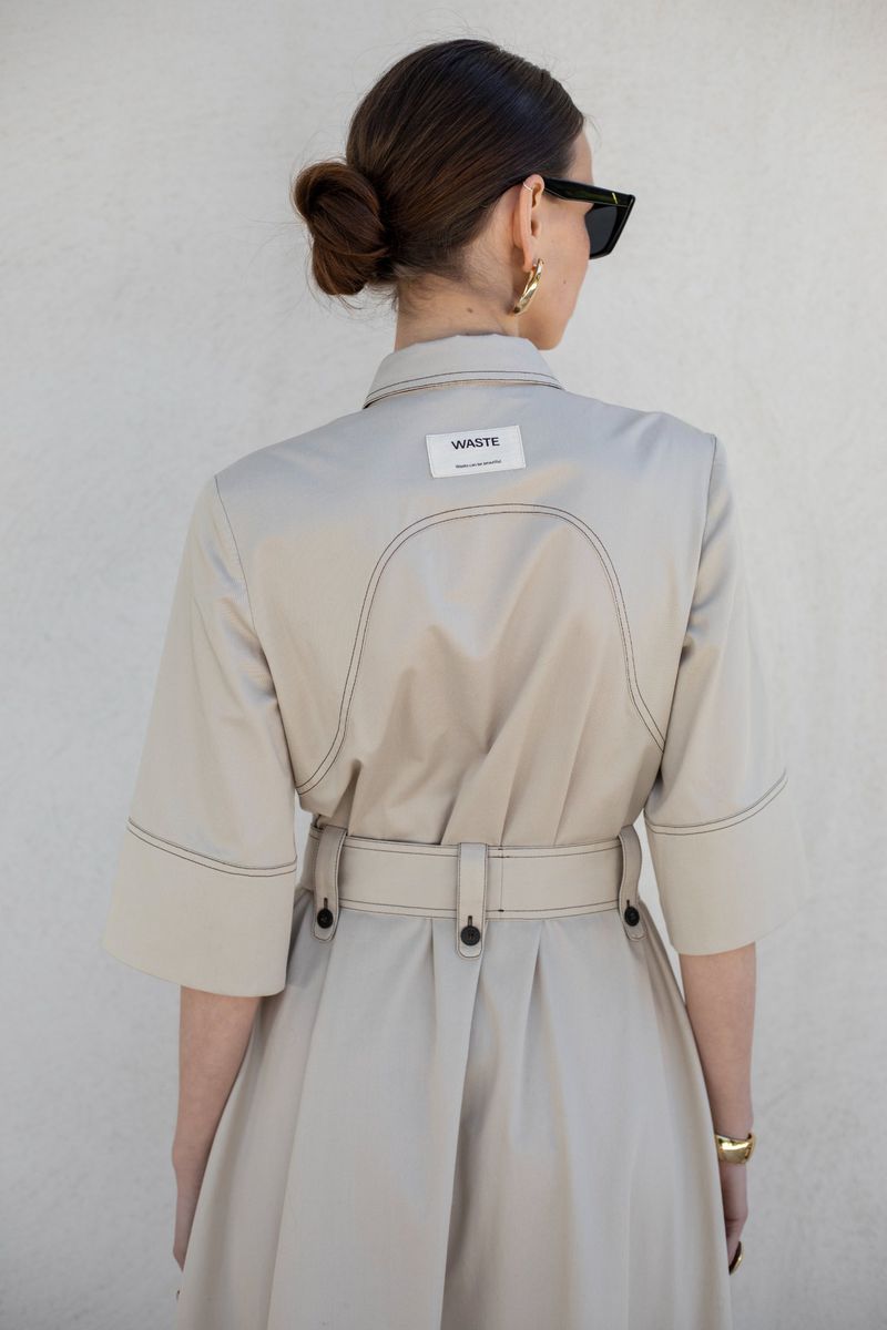 Back view of a woman in a tan trench coat dress, wearing sunglasses and gold hoop earrings, standing against a neutral wall.