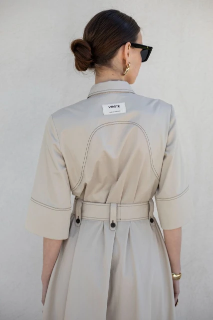Back view of a woman in a tan trench coat dress, wearing sunglasses and gold hoop earrings, standing against a neutral wall.