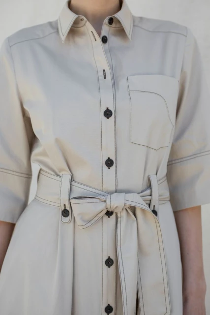 Woman in beige shirt dress with buttons and matching belt tied in bow, front pocket, and contrast stitching details.