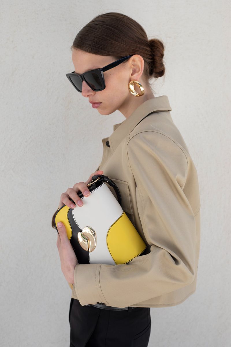 Model wearing black sunglasses, a beige jacket, and gold earrings while holding a yellow and white colorblock handbag.