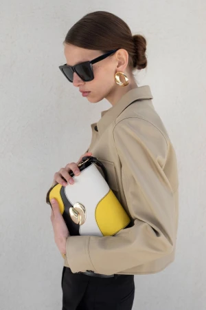 Model wearing black sunglasses, a beige jacket, and gold earrings while holding a yellow and white colorblock handbag.