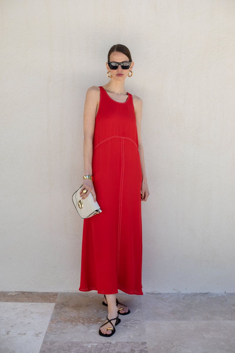 A woman in a red dress, sunglasses, and gold earrings stands against a neutral wall holding a white clutch, creating a fashion-forward portrait.