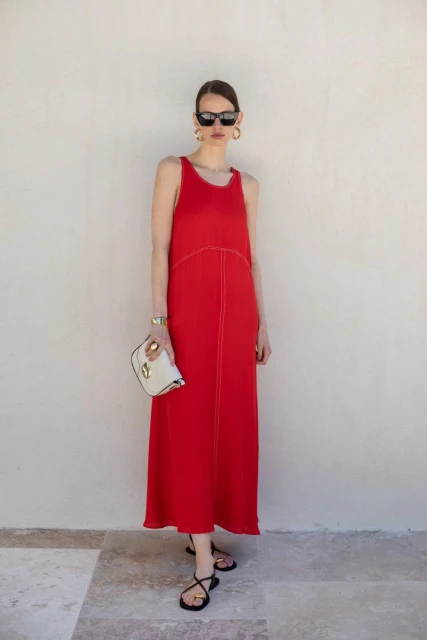 A woman in a red dress, sunglasses, and gold earrings stands against a neutral wall holding a white clutch, creating a fashion-forward portrait.