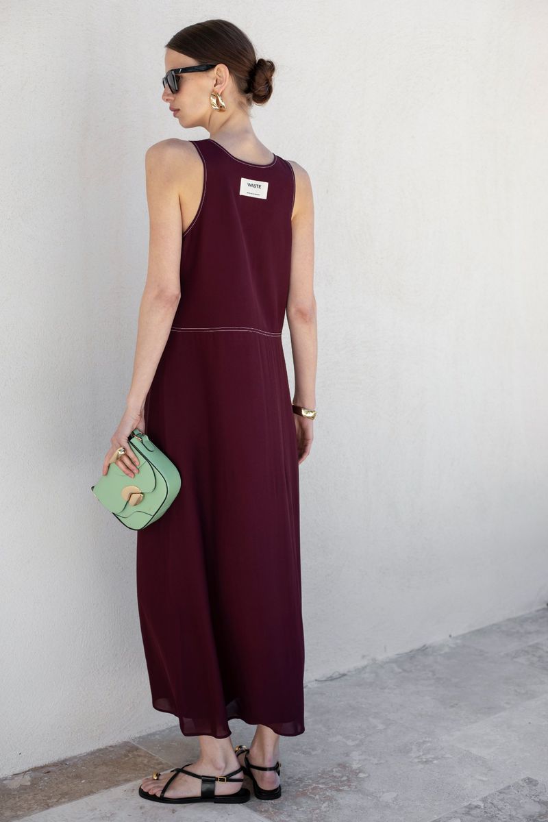 Woman in a burgundy dress and black sandals, carrying a mint green purse, stands outdoors against a white wall in a fashion shot.