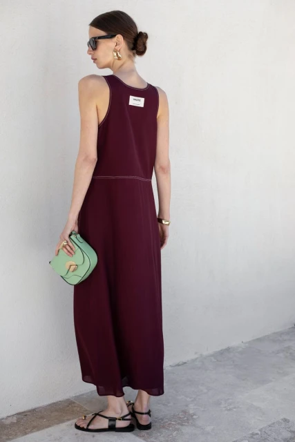 Woman in a burgundy dress and black sandals, carrying a mint green purse, stands outdoors against a white wall in a fashion shot.