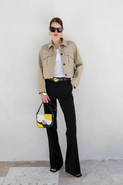 Full-length shot of an adult woman in a beige cropped jacket, white top, black pants, sunglasses, and accessories, holding a purse.