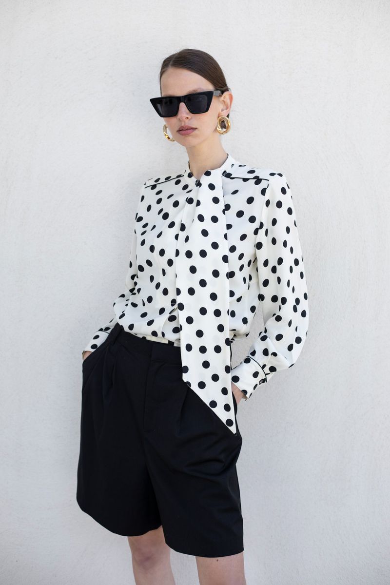 Woman in a polka dot blouse and black shorts stands against a white wall, accessorized with sunglasses and gold earrings, fashion pose.