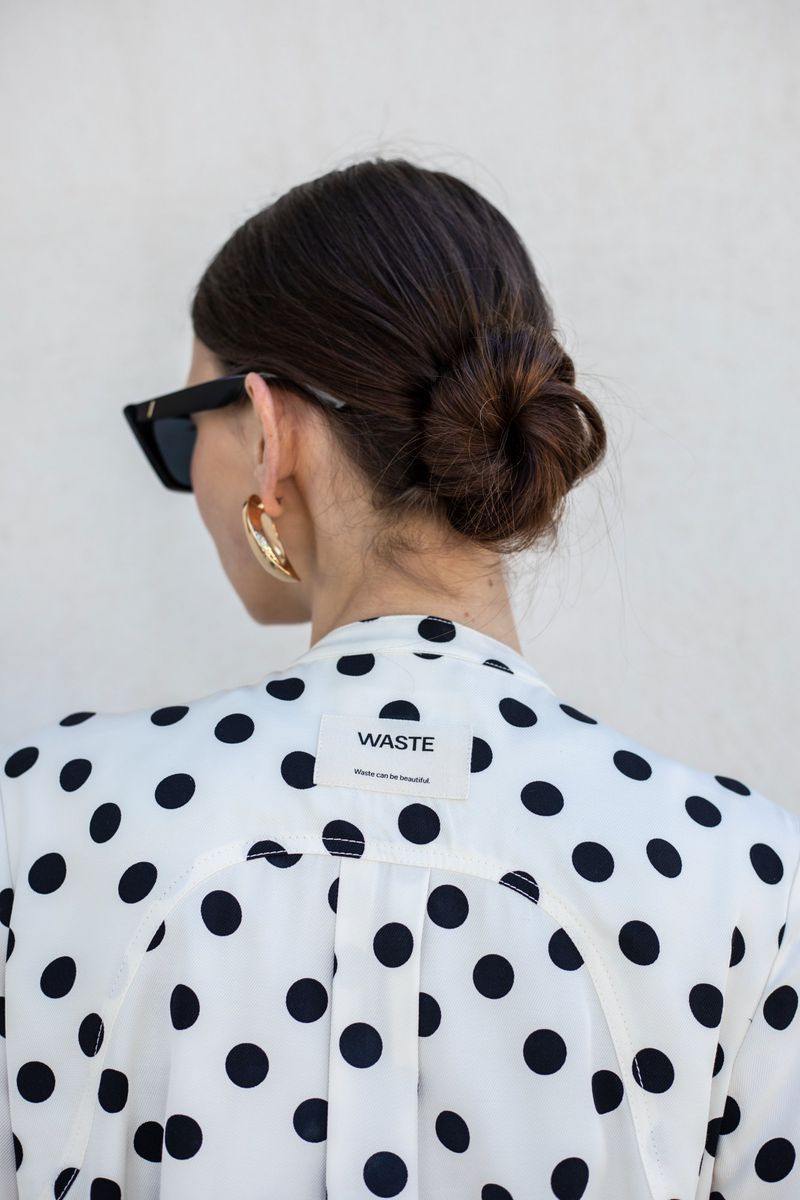 Woman with dark hair styled in a bun, seen from the back, wearing a white polka dot blouse, gold hoop earrings, and sunglasses.