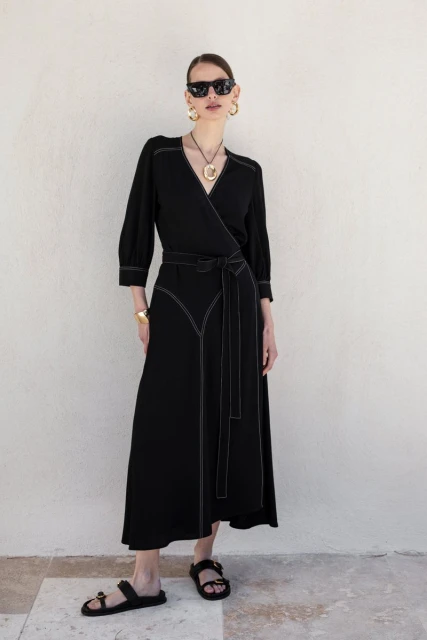 An adult female poses against a neutral wall wearing a black wrap dress with white stitching, complemented by black sandals and gold jewelry.