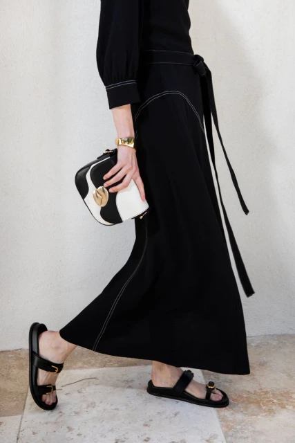 Woman in black dress walks indoors holding a black and white purse, accessorized with a gold bracelet and wearing black sandals.