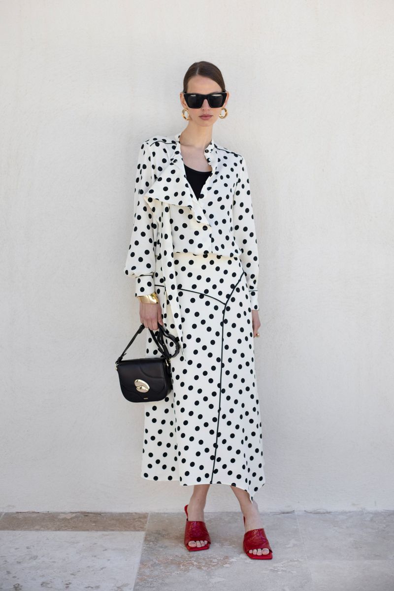 Elegant adult female in polka-dot dress, sunglasses, and red sandals poses against a white wall with a stylish black purse.