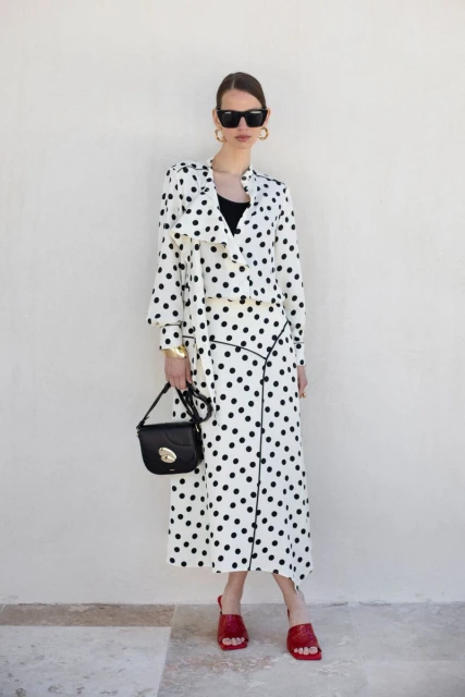 Elegant adult female in polka-dot dress, sunglasses, and red sandals poses against a white wall with a stylish black purse.
