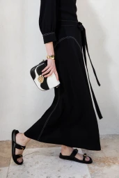 Model in a black maxi skirt with white contrast stitching, holding a black and white handbag and wearing flat black sandals.