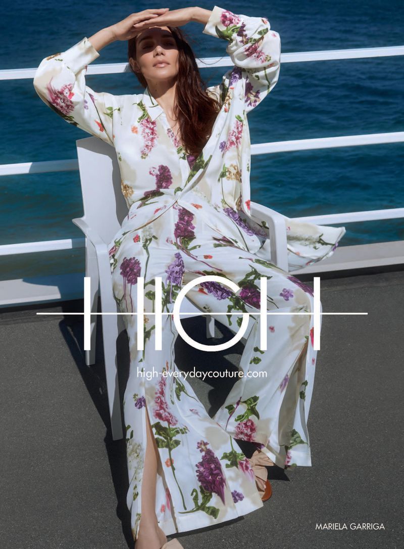 A woman in floral pajamas sits on a white chair, hands above her brow, blocking the sun, with ocean in the background.