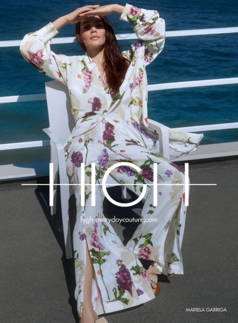 A woman in floral pajamas sits on a white chair, hands above her brow, blocking the sun, with ocean in the background.