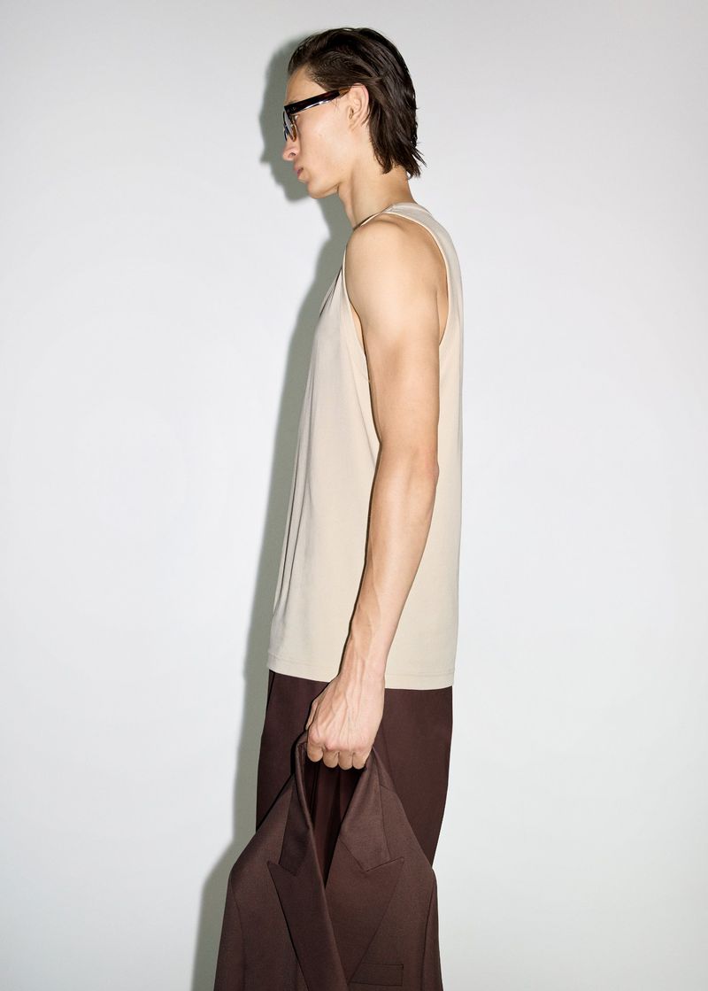 Studio shot of a slender adult male in a beige tank top and brown pants, holding a brown jacket against a plain white background.