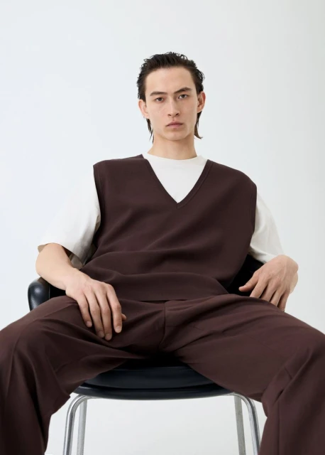 A stylish adult man in a brown vest and matching pants sits on a black stool against a bright white background, looking at the camera.