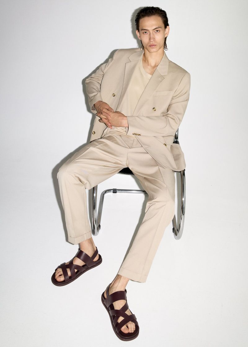 An adult male model sits on a metal chair in a photo studio wearing a tan suit, t-shirt, and brown leather sandals, against a white backdrop.