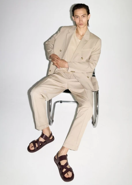 An adult male model sits on a metal chair in a photo studio wearing a tan suit, t-shirt, and brown leather sandals, against a white backdrop.