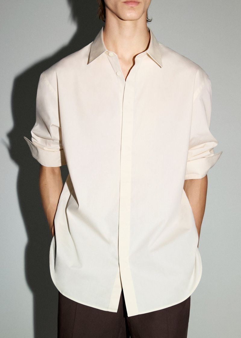 Man wearing an open beige button-down shirt with sleeves rolled up and brown pants in a studio with a soft gray backdrop.