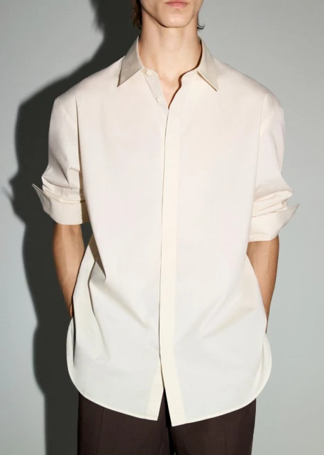 Man wearing an open beige button-down shirt with sleeves rolled up and brown pants in a studio with a soft gray backdrop.