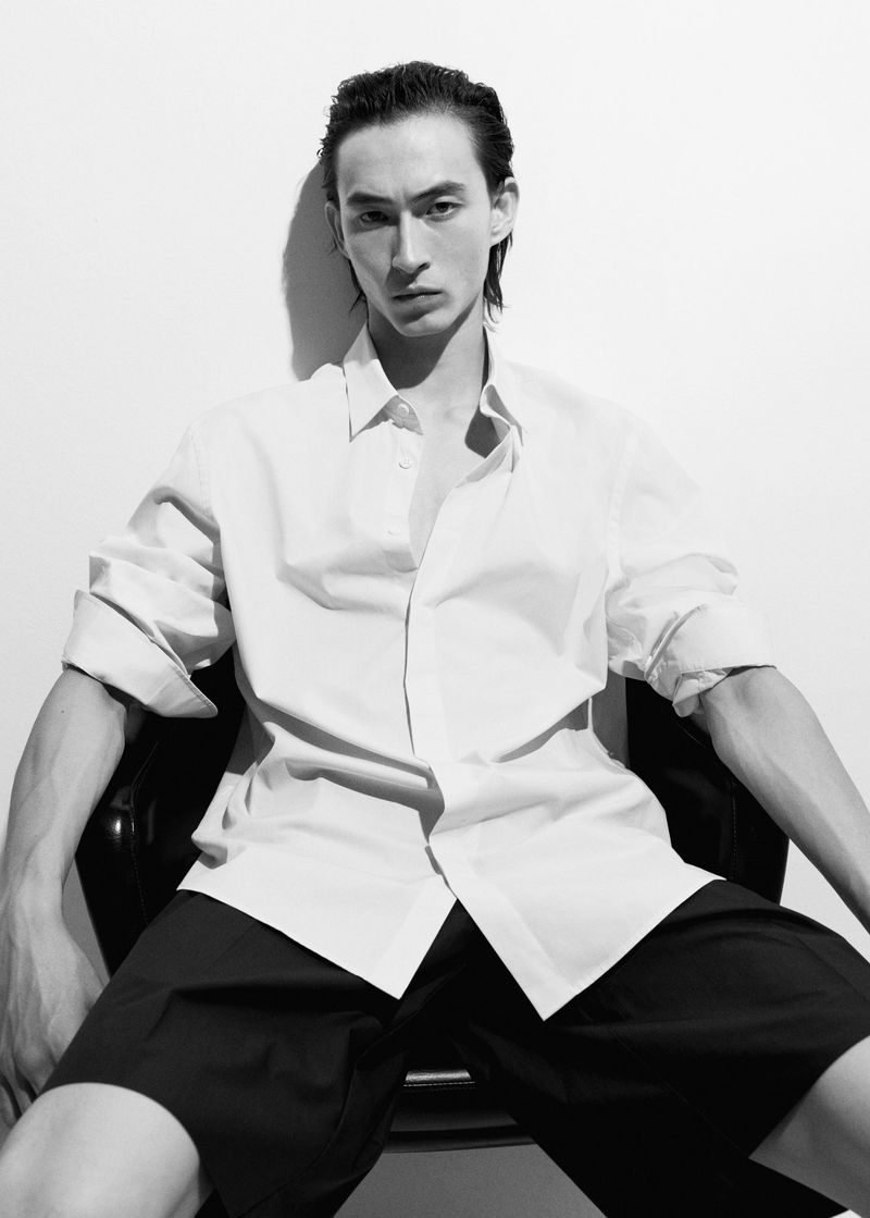 Monochrome portrait of an adult male model with slicked-back hair, wearing an unbuttoned white shirt and black shorts, seated indoors.