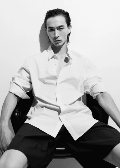 Monochrome portrait of an adult male model with slicked-back hair, wearing an unbuttoned white shirt and black shorts, seated indoors.