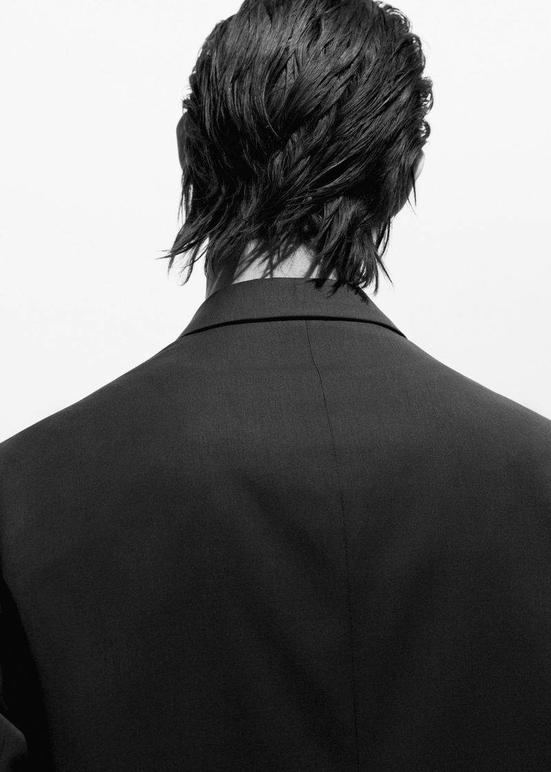 Black-and-white studio portrait from behind of a person with dark, wet hair wearing a black suit jacket against a white backdrop.
