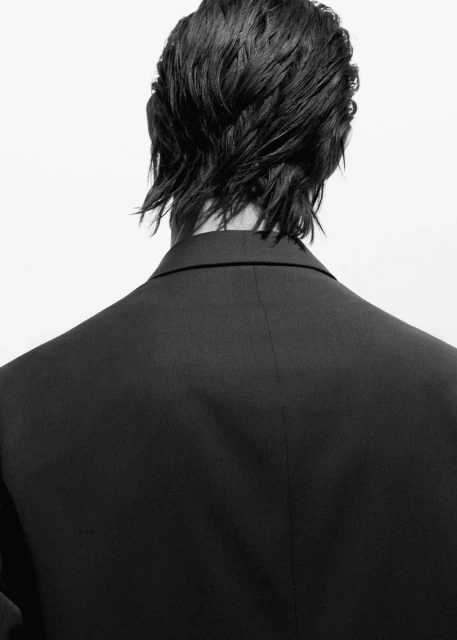 Black-and-white studio portrait from behind of a person with dark, wet hair wearing a black suit jacket against a white backdrop.