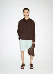 Full-length portrait of a male model wearing a dark brown jacket, light blue shorts, and sandals against a white studio background.