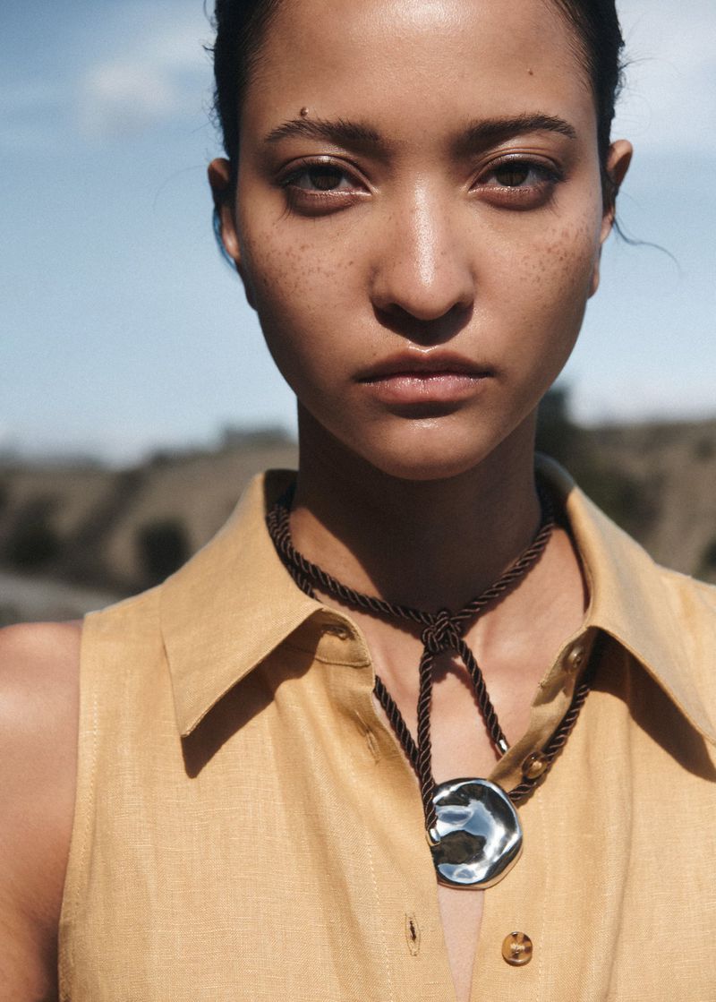 Close-up of a woman with freckles wearing a light-yellow dress and silver pendant necklace in an outdoor setting with natural light.