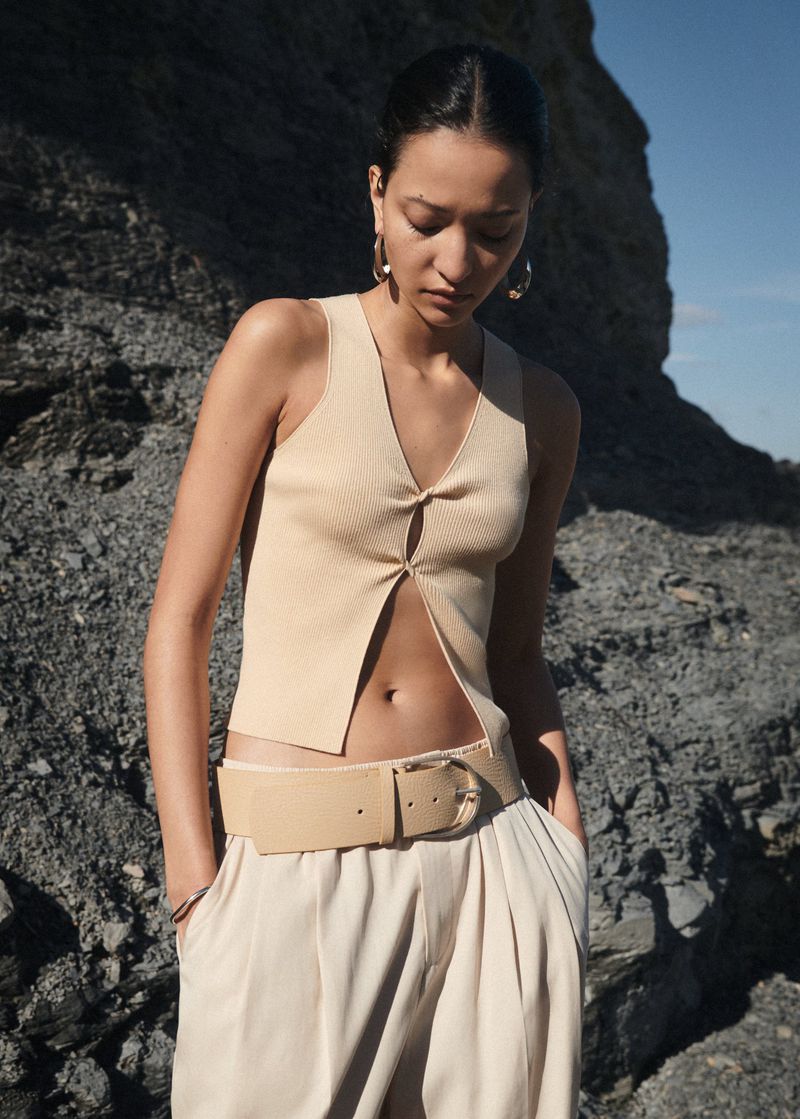 A woman models beige knit top, wide belt, and baggy pants outdoors. The neutral-toned fashion outfit complements the rocky landscape.