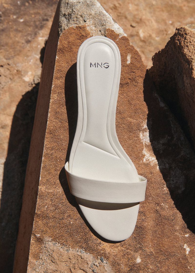 Cream-colored sandal with a single strap is displayed on a textured brown rock surface, with the brand MNG visible on the insole.