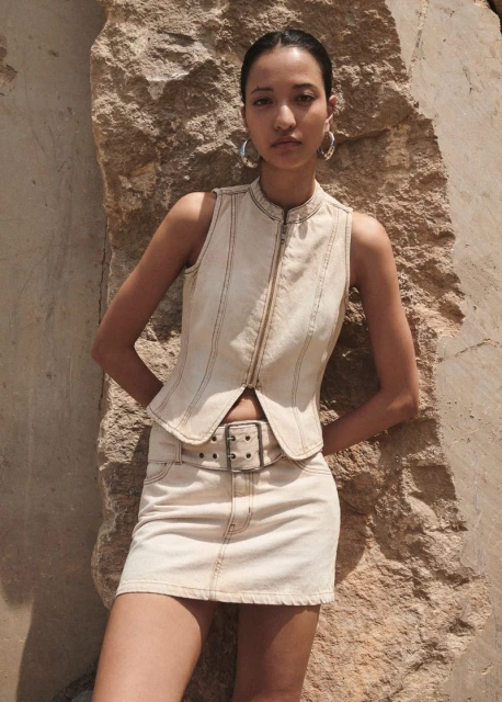 Adult female poses outdoors against a textured rock wearing a denim vest and mini-skirt with large hoop earrings, looking at the camera.