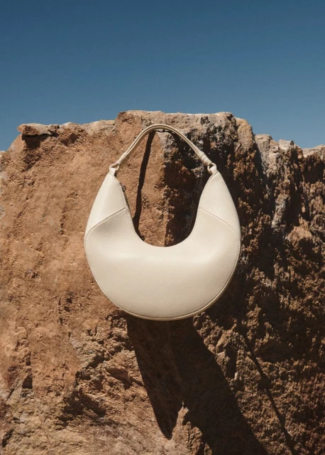 Fashion product shot of a crescent-shaped cream-colored shoulder bag against a textured rock and a clear blue sky backdrop.