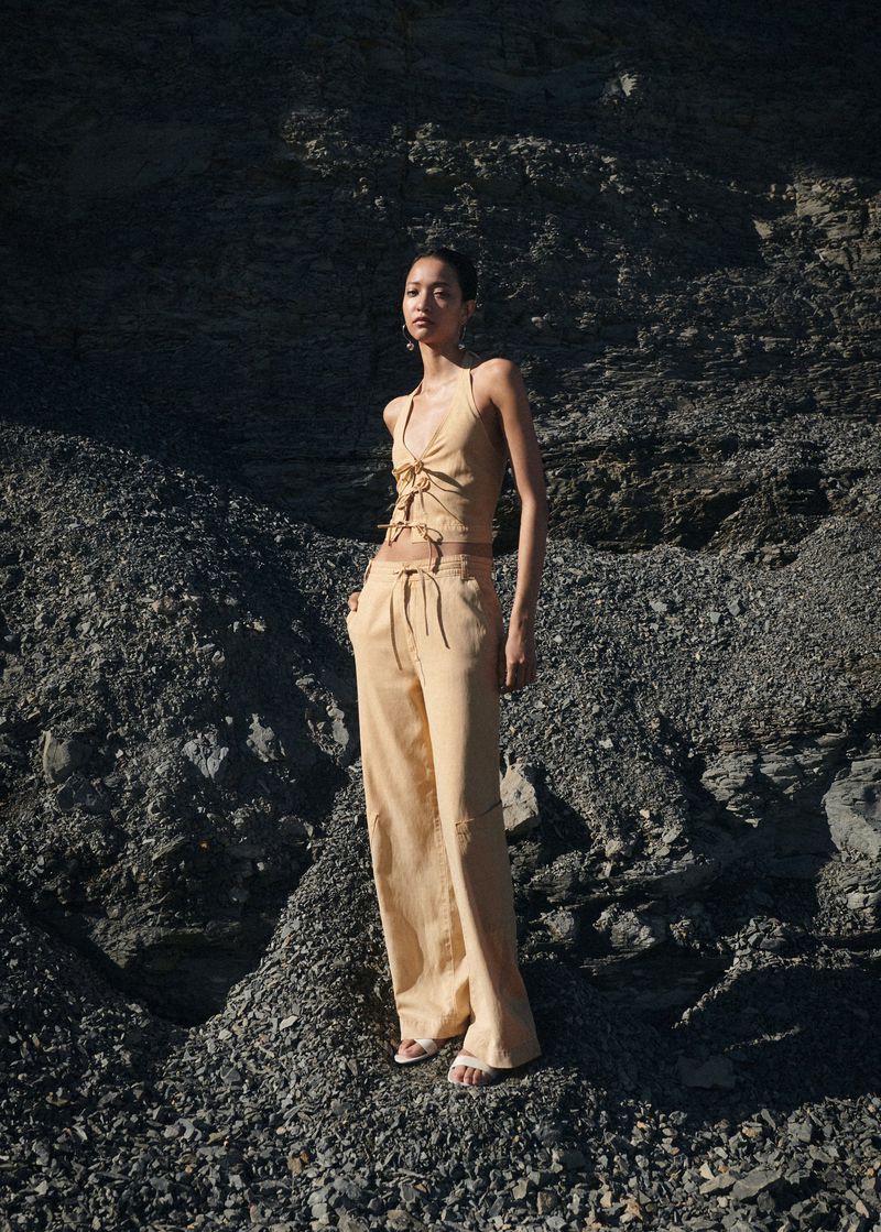 A woman in a beige halter top and pants stands on a rocky surface in a fashion shot, styled with neutral colors and minimal accessories.