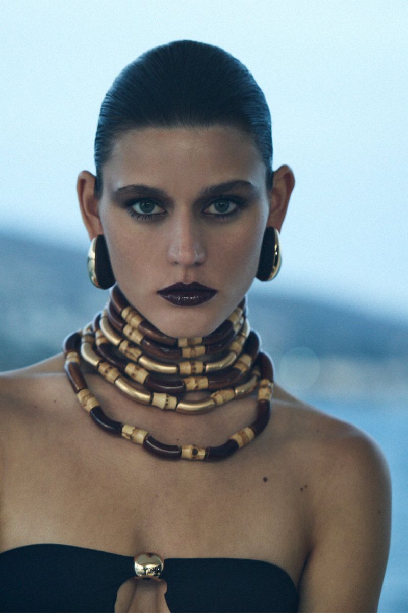 Woman wearing layered wooden tube necklace and gold hoop earrings in a fashion headshot against a blurred blue background.
