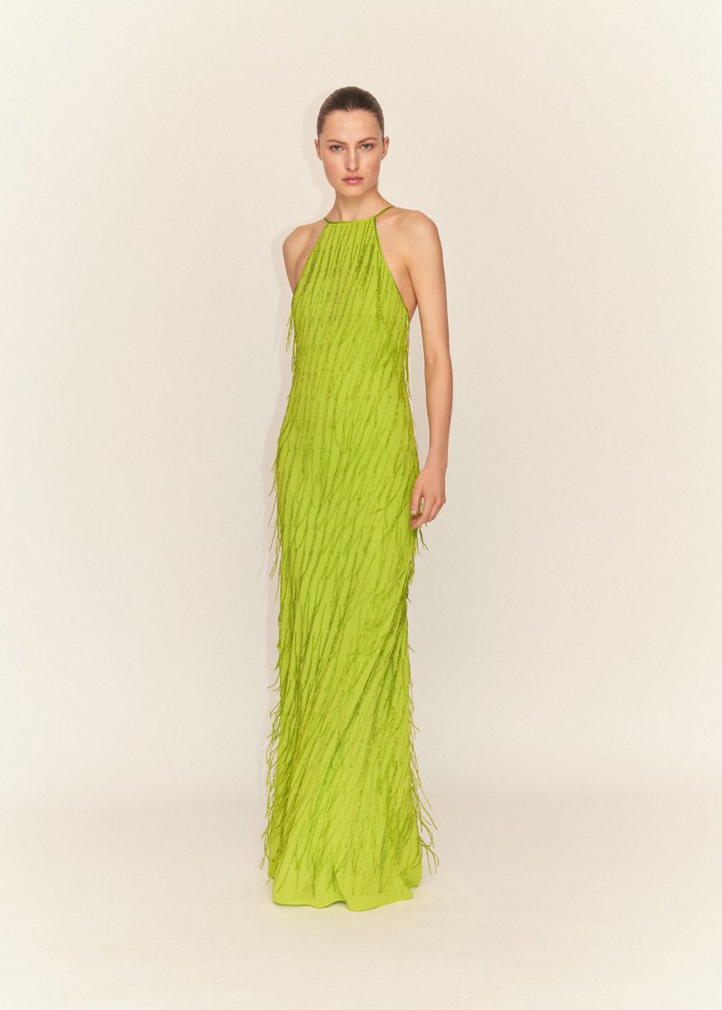Elegant woman poses in full-length shot, wearing a fringed, light green halter gown against a neutral backdrop in a fashion editorial setting.