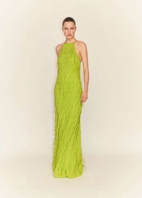 Elegant woman poses in full-length shot, wearing a fringed, light green halter gown against a neutral backdrop in a fashion editorial setting.