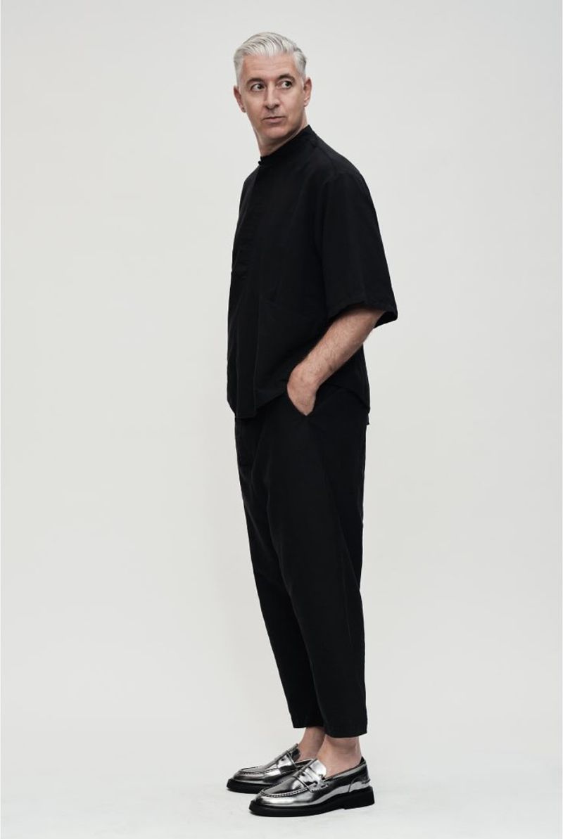 Studio shot of a mature man wearing black clothing and shiny silver loafers, standing in profile on a seamless white backdrop.