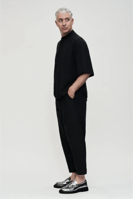 Studio shot of a mature man wearing black clothing and shiny silver loafers, standing in profile on a seamless white backdrop.