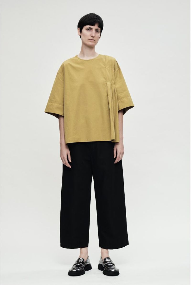 Full-length shot of a woman in a loose-fitting gold blouse, black pants, and black shoes, posing against a neutral background.