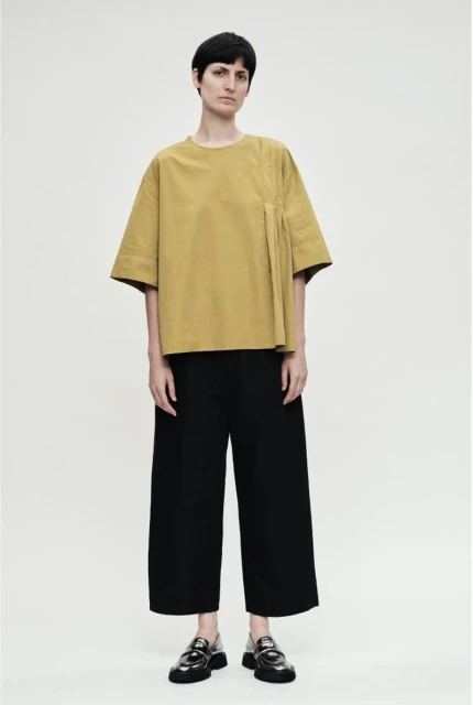 Full-length shot of a woman in a loose-fitting gold blouse, black pants, and black shoes, posing against a neutral background.